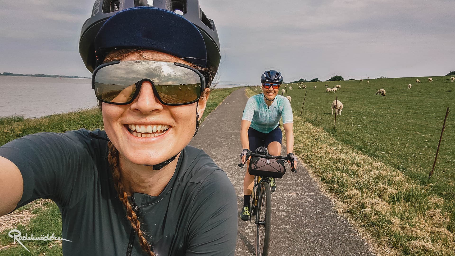 Frau lcht in die Kamera mit Helm und Fahrradbrille, eine weitere Frau fährt auf dem Fahrrad hinter ihr