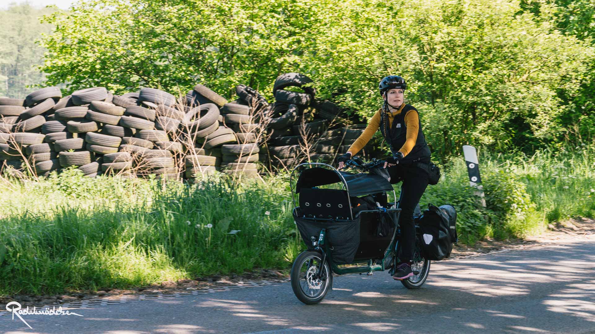 Frau auf dem muli Kompakt-Lastenrad radelt auf der Straße, im Hintergrund Reifenstapel und Natur